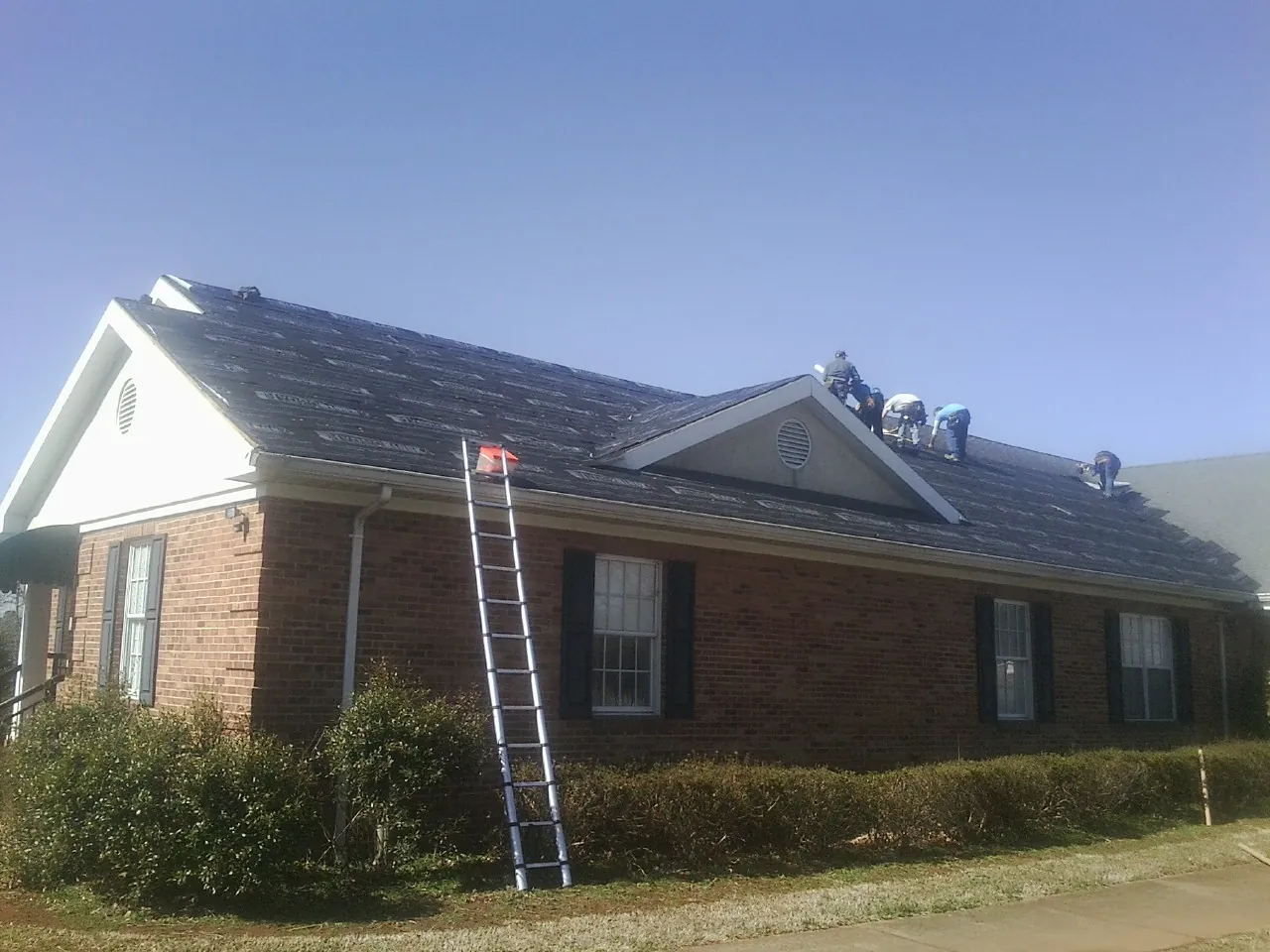 Crew tearing shingles off a brick ranch roof with a ladder set — Emergency Roof Repair work by our crew in Miller Place